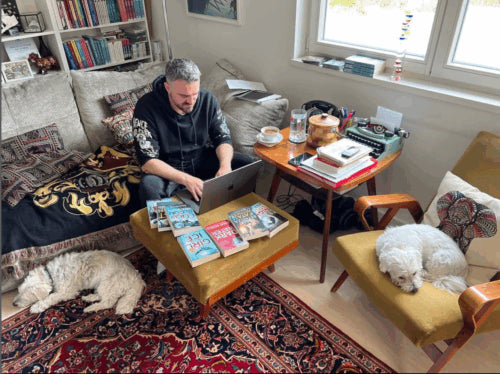 Robert Bryndza author working on a laptop in a cozy living room with two dogs and books around.